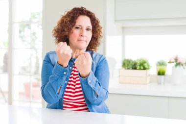 Evde kot ceket giyen kıvırcık saçlı orta yaş yaşlı kadın yumruk savunma jesti ile mücadeleye hazır, kızgın ve üzgün yüz, sorun korkuyor