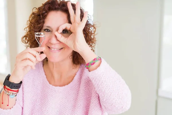 Senior woman using eyelashes curler and mascara with happy face smiling doing ok sign with hand on eye looking through fingers