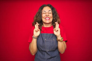 Kırmızı izole arka plan gesturing parmak üzerinde önlük üniforma giyen orta yaş yaşlı kadın umut ve gözleri kapalı gülümseyerek geçti. Şans ve batıl kavram.