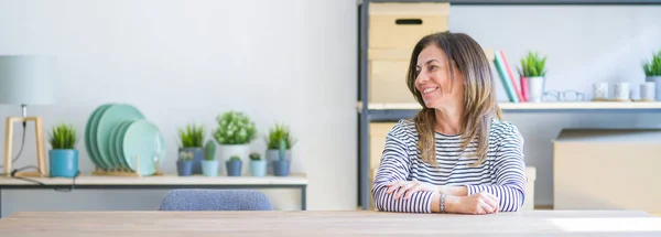 Wide angle photo of middle age senior woman sitting at the table at home looking away to side with smile on face, natural expression. Laughing confident.