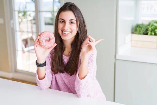 ピンクのチョコチップを食べる美しい若い女性ドーナツは 手と指を横に指差して非常に幸せ — ストック写真