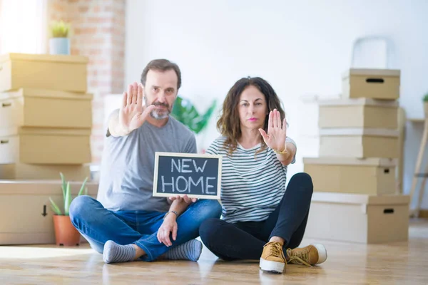 Middle age senior couple sitting on the floor holding blackboard moving to a new home with open hand doing stop sign with serious and confident expression, defense gesture
