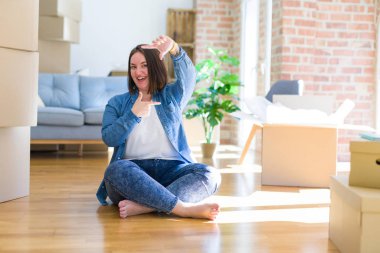 Young plus size woman sitting on the floor around cardboard boxes moving to a new home smiling making frame with hands and fingers with happy face. Creativity and photography concept.