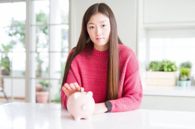 Güzel Asyalı kadın ciddi düşünme akıllı yüz üzerinde kendine güvenen bir ifade ile piggy çanta içinde bir sikke koyarak