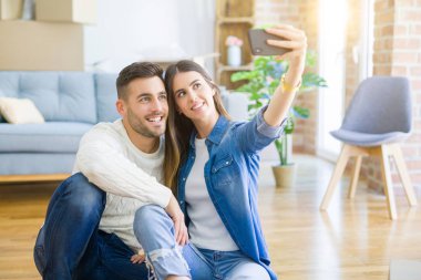 Yeni evin zemininde oturup akıllı telefon kullanarak selfie fotoğrafı çeken genç çift, yeni daireye taşındığı için mutlu bir şekilde gülümsüyor