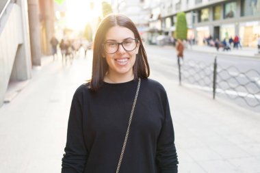 Şehir sokaklarında yürürken heyecanlı gülümseyen güzel genç esmer kadın, şehirde açık havada mutlu ve güven ifade ayakta