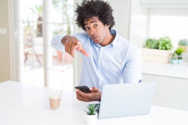 African American business man using smartphone and laptop pointing with finger to the camera and to you, hand sign, positive and confident gesture from the front