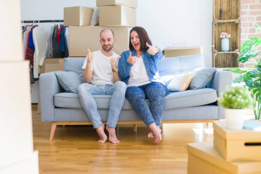 Young couple sitting on the sofa arround cardboard boxes moving to a new house success sign doing positive gesture with hand, thumbs up smiling and happy. Cheerful expression and winner gesture.