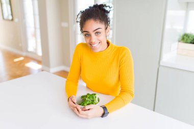 Young african american girl eating heatlhy green broccoli with a happy face standing and smiling with a confident smile showing teeth