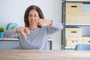 Orta yaş yaşlı kadın evde masada oturan başparmak yukarı ve aşağı yapıyor, anlaşmazlık ve anlaşma ifade. Çılgın çatışma