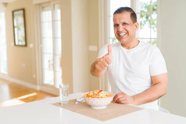 Middle age man eating rice at home doing happy thumbs up gesture with hand. Approving expression looking at the camera with showing success.