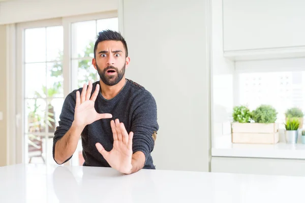 Handsome hispanic man wearing casual sweater at home afraid and terrified with fear expression stop gesture with hands, shouting in shock. Panic concept.