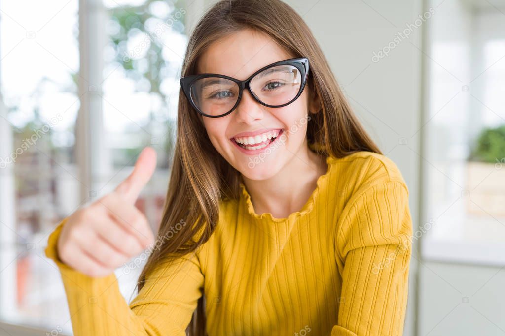 Beautiful young girl kid wearing glasses doing happy thumbs up gesture with hand. Approving expression looking at the camera showing success.