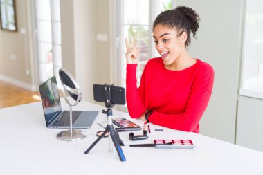 Young african american influencer woman recording make up tutorial with a big smile on face, pointing with hand and finger to the side looking at the camera.