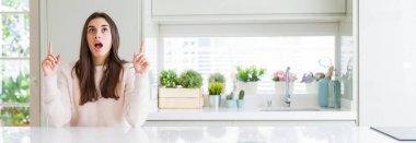 Güzel genç kadın evde beyaz masaya oturan geniş açı resim hayret ve yukarıya bakıp parmakları ile işaret şaşırttı ve silah kaldırdı.