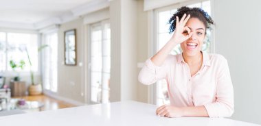 Wide angle of beautiful african american woman with afro hair doing ok gesture with hand smiling, eye looking through fingers with happy face.