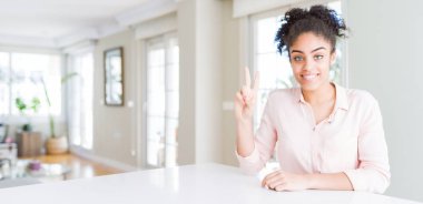 Wide angle of beautiful african american woman with afro hair showing and pointing up with fingers number two while smiling confident and happy.