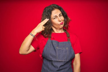 Orta yaş yaşlı kadın kırmızı izole arka plan üzerinde önlük üniforma giyen Çekim ve kendini silah gibi baş el ve parmakları işaret öldürme, intihar jesti.