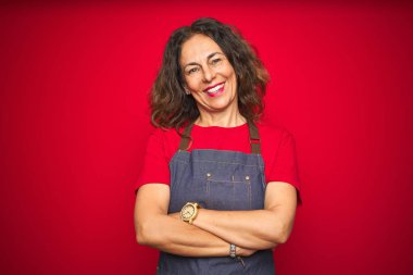 Kırmızı izole arka plan üzerinde önlük üniforma giyen orta yaş yaşlı kadın kamera bakarak çapraz kolları ile gülümseyen mutlu yüz. Pozitif kişi.