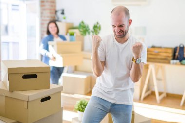 Young couple arround cardboard boxes moving to a new house, bald man standing at home very happy and excited doing winner gesture with arms raised, smiling and screaming for success. Celebration concept.