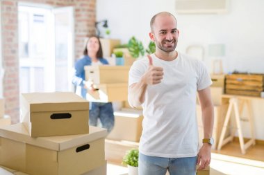 Young couple arround cardboard boxes moving to a new house, bald man standing at home doing happy thumbs up gesture with hand. Approving expression looking at the camera with showing success.