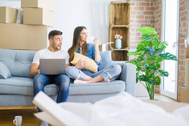Young beautiful couple relaxing sitting on the sofa of new house using computer laptop pointing and showing with thumb up to the side with happy face smiling