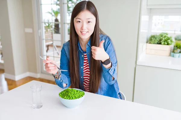 Beautiful Asian woman eating fresh green peas happy with big smile doing ok sign, thumb up with fingers, excellent sign