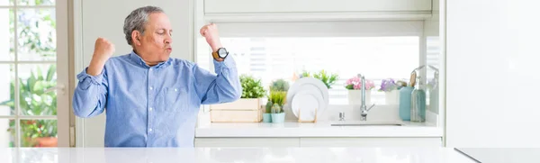 Wide angle perspective of handsome senior man at home showing arms muscles smiling proud. Fitness concept.