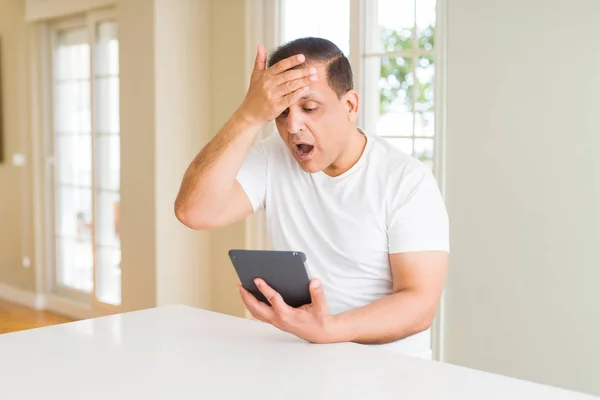 Middle age man using digital table at home stressed with hand on head, shocked with shame and surprise face, angry and frustrated. Fear and upset for mistake.