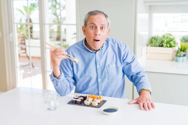 Handsome senior man eating take away sushi using chopsticks at home scared in shock with a surprise face, afraid and excited with fear expression