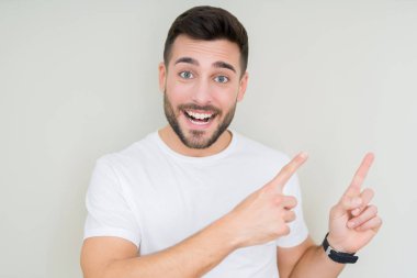 Gülümseyen ve iki elleri ve parmakları ile tarafına işaret kamera bakarak izole arka plan üzerinde rahat beyaz t-shirt giyen genç yakışıklı adam.