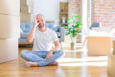 Genç kel adam karton kutuların etrafında oturmuş yeni evine taşınıyor. El hareketleriyle gülümsüyor, gözleriyle mutlu yüzle parmaklarının arasından bakıyor..