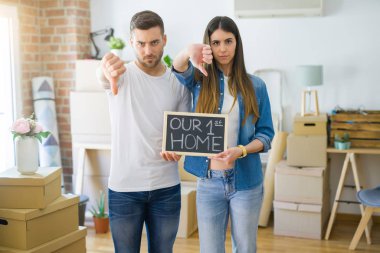 Young beautiful couple holding blackboard with our first home text at new house with angry face, negative sign showing dislike with thumbs down, rejection concept