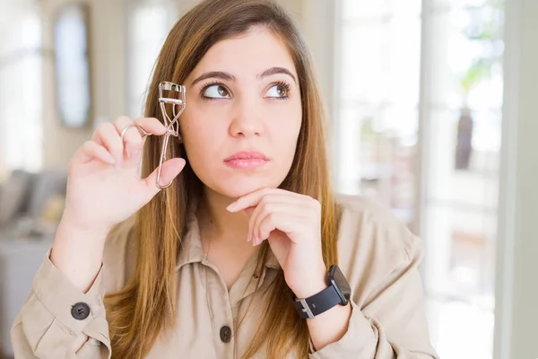 Beautiful young woman curling eyelashes using eyelash curler serious face thinking about question, very confused idea