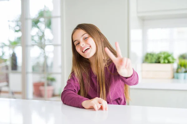 美丽的年轻女孩在白色的桌子上微笑着看着相机显示手指做胜利的标志 — 图库照片