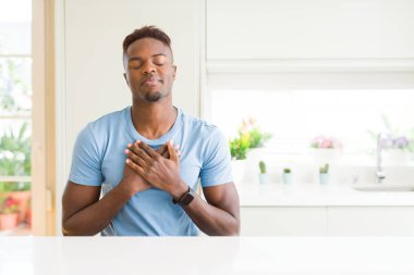 Evde rahat t-shirt giyen yakışıklı Afrikalı Amerikalı adam yüzünde kapalı gözler ve minnettar jest ile göğsünde eller ile gülümsüyor. Sağlık konsepti.