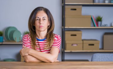 Orta yaş yaşlı kadın evde masada oturan şüpheci ve sinirli, çapraz kolları ile yüzünde ifade onaylamayan. Negatif kişi.