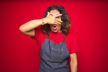 Kırmızı izole arka plan üzerinde önlük giyen orta yaş yaşlı kadın şok yüz ve gözleri el ile kaplayan gözetleme, utanmış ifade ile parmaklardan bakarak.