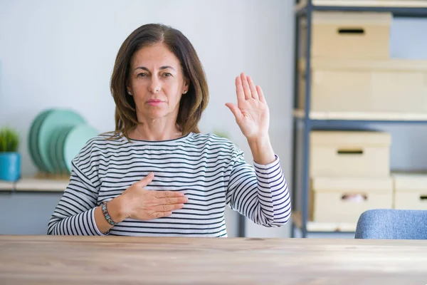 中年老妇人坐在家里的餐桌上 手放在胸前 张开手掌 做出忠诚誓言 — 图库照片