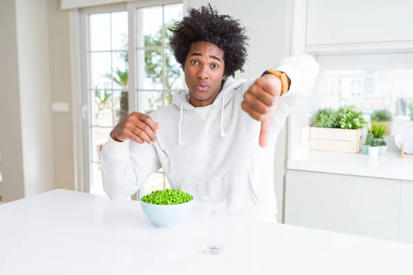 African American man eating fresh green peas at home with angry face, negative sign showing dislike with thumbs down, rejection concept