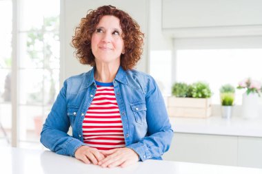 Kıvırcık saçlı orta yaş yaşlı kadın evde kot ceket giyen yan bakarak ve düşünme uzağa bakıyor.