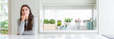 Çene düşünme hakkında soru, dalgın ifade üzerine el ile evde beyaz masaya oturan güzel genç kadın geniş açı resim. Düşünceli yüzüyle gülümsüyor. Şüphe kavramı.