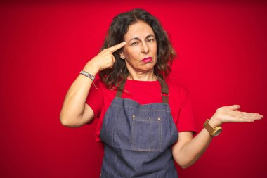 Kırmızı izole arka plan üzerinde önlük üniforma giyen orta yaş yaşlı kadın karışık ve açık avuç içi ile rahatsız kopya alanı gösteren ve alnına parmak işaret. Bir düşün..
