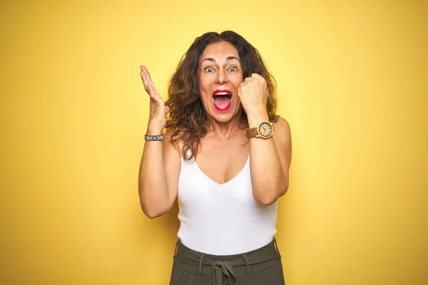 Middle age senior woman showing wrist watch over yellow isolated background very happy and excited, winner expression celebrating victory screaming with big smile and raised hands