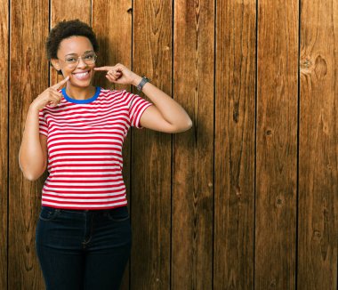 Kendinden emin gösterilen ve işaret parmakları diş ve ağız ile gülümseyen izole arka plan üzerinde gözlük güzel genç Afro-Amerikan kadın. Sağlık kavramı.