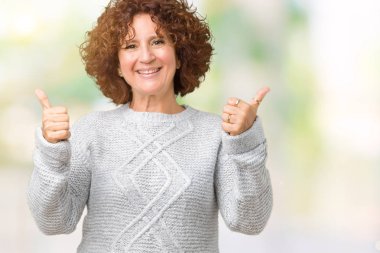 El ile pozitif jest yapıyor izole arka plan başarı işareti üzerinde kış kazak giyen güzel orta ager üst düzey kadın başparmak gülümseyen ve mutlu. Neşeli bir ifadeyle, kazanan jest kameraya bakıyor.