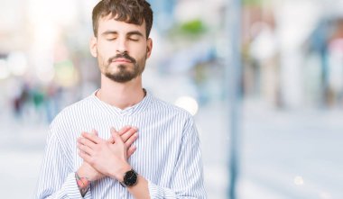 Genç yakışıklı iş adamı izole arka plan ile gülümseyen üzerinde kapalı gözler ve yüzünde minnettar jest ile göğüs üzerinde eller. Sağlık kavramı.