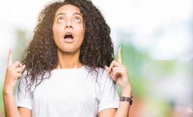 Kıvırcık saçlı rahat beyaz t-shirt hayran ve görünümlü ve parmaklar ve yükseltilmiş kollar ile işaret şaşırttı giyen genç güzel kız.