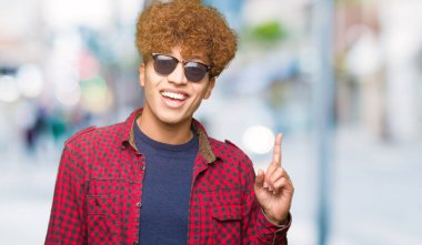 Yakışıklı genç afro saçlı başarılı fikirle parmak işaret güneş gözlüğü takıyor. Çıktıktan ve mutlu. Bir numara.