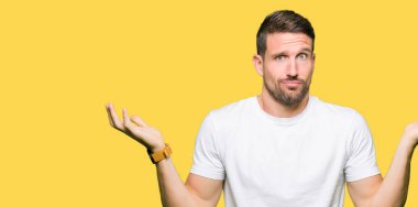 Yakışıklı adam rahat beyaz t-shirt clueless ve şaşkın bir ifadeyle kollar ve eller kaldırdı giyiyor. Şüphe kavramı.